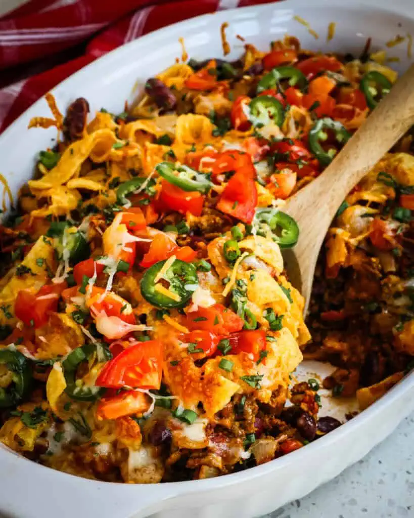 A wooden spoon full of frito casserole over a casserole dish full of frito pie. 