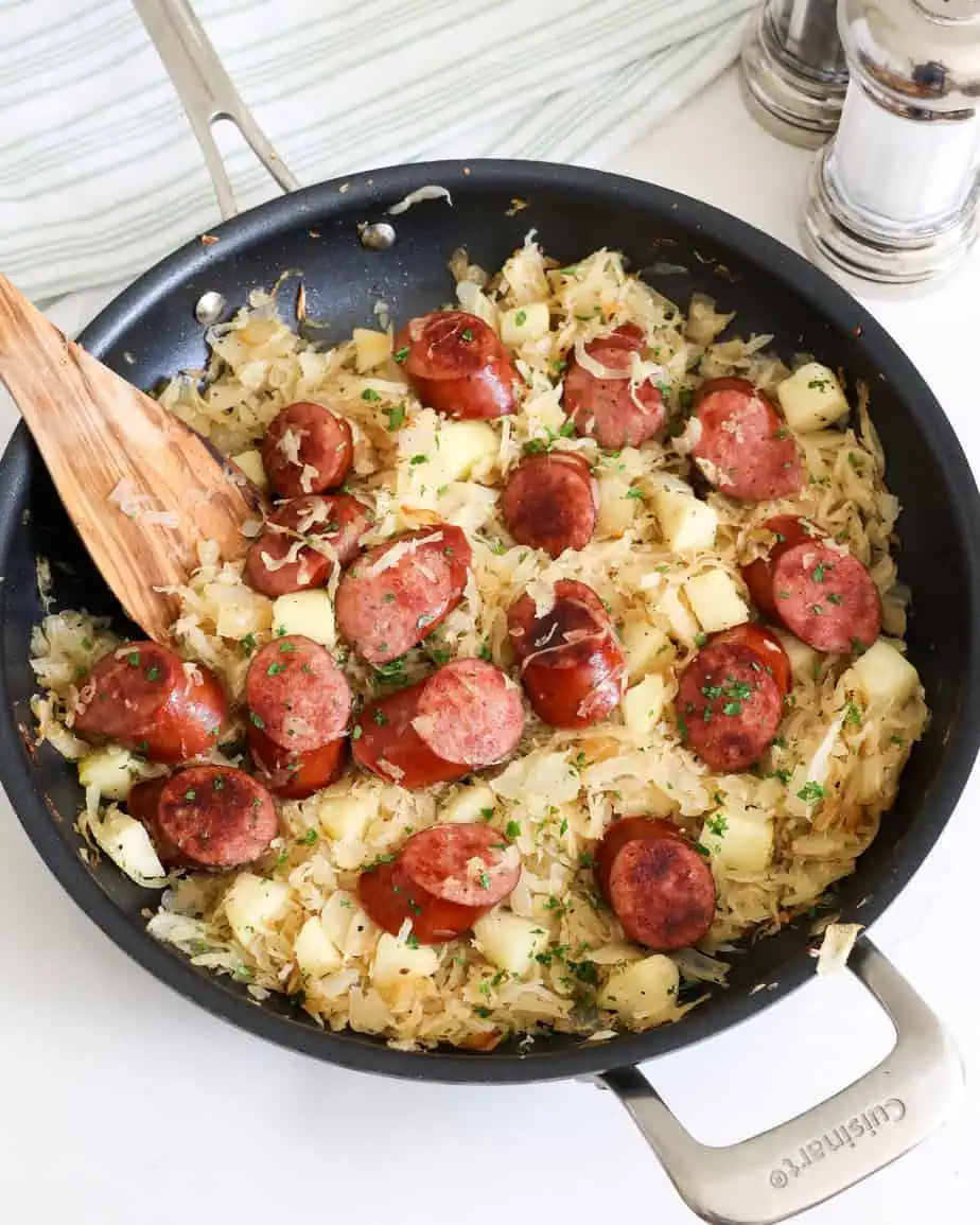 Polska kielbasa, sauerkraut and apples in a skillet with a wooden spatula and a salt and pepper shaker off to the side. 