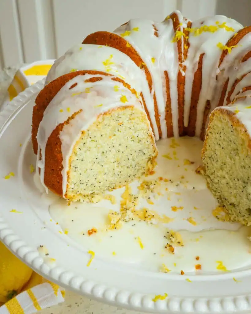 Lemon poppy seed cake with a lemon glaze on a cake stand with a couple of slices cut out of it. 