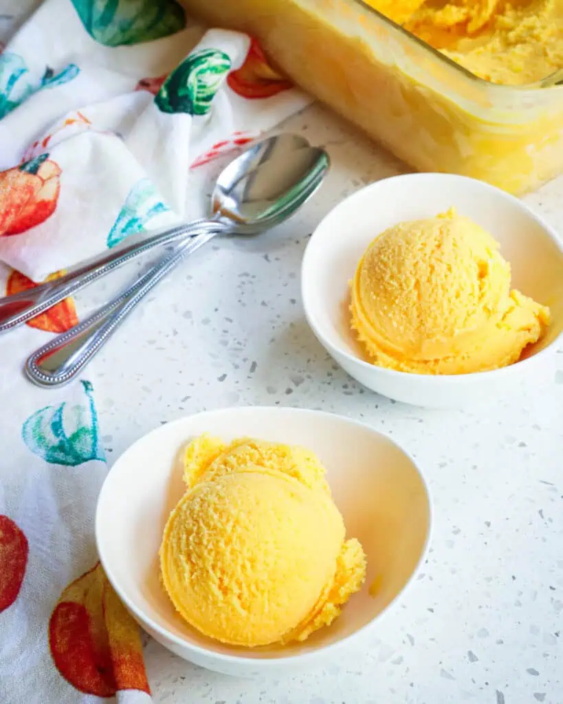 Two bowls with a large scoop of orange sherbet in a bowl with two spoons off to the side. 