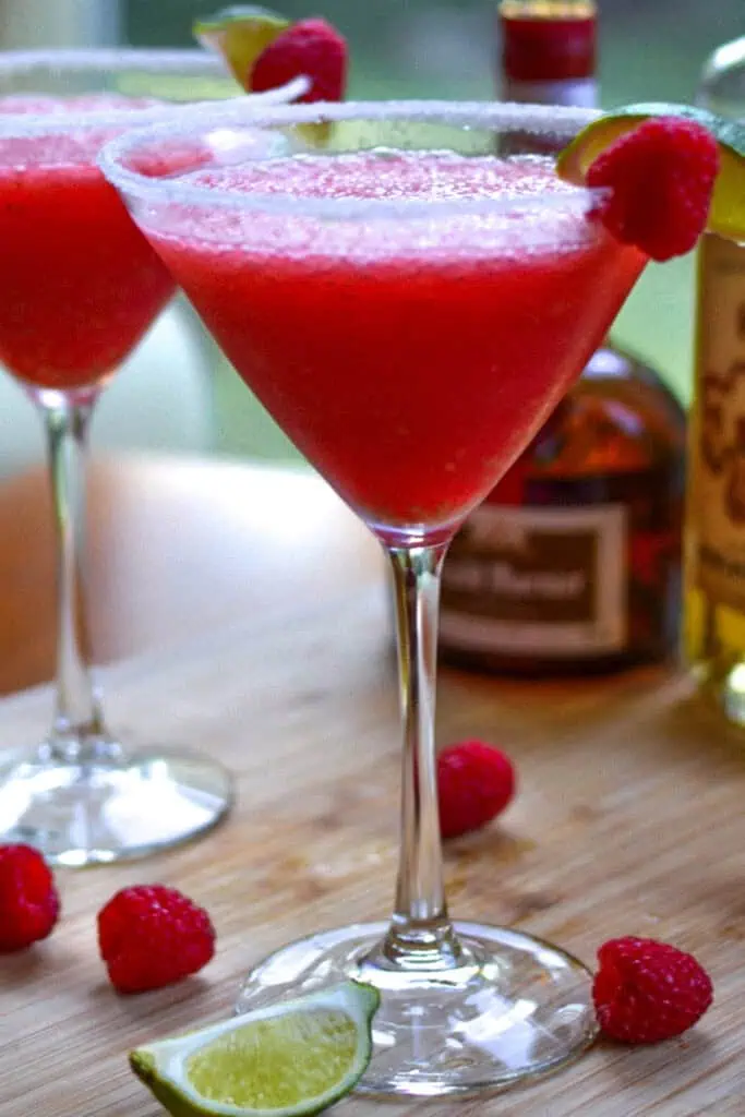 A raspberry margarita garnished with a lime slice and a raspberry with a botle of grand marnier in the background. 