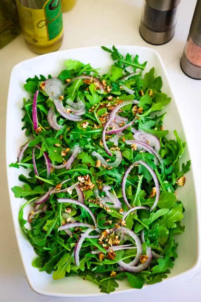 A bowl full of arugula lettuce with thin slices of red onion and chopped pecan. 