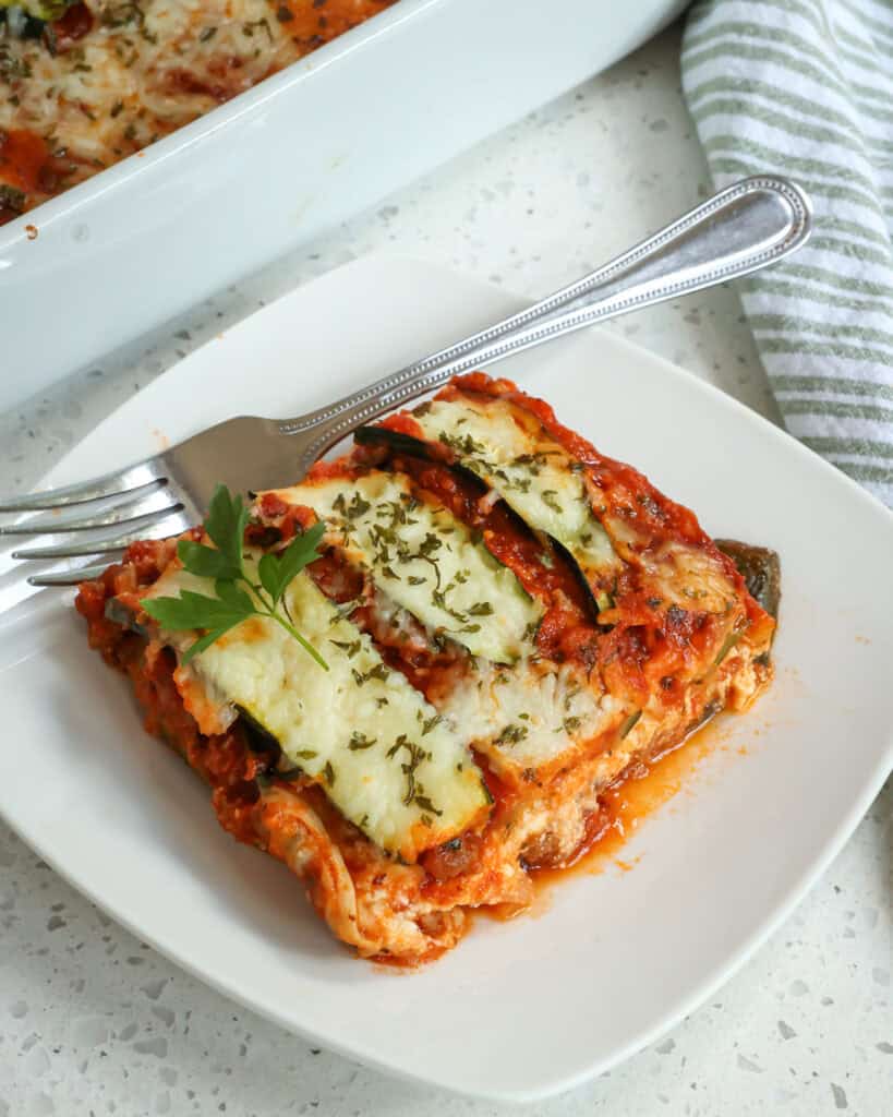 Zucchini lasagna on a plate with a fork and a sprig of parsley. 