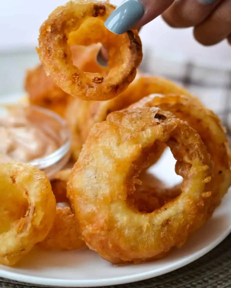 A lady holding up a beer battered onion ring over a plate of them. 