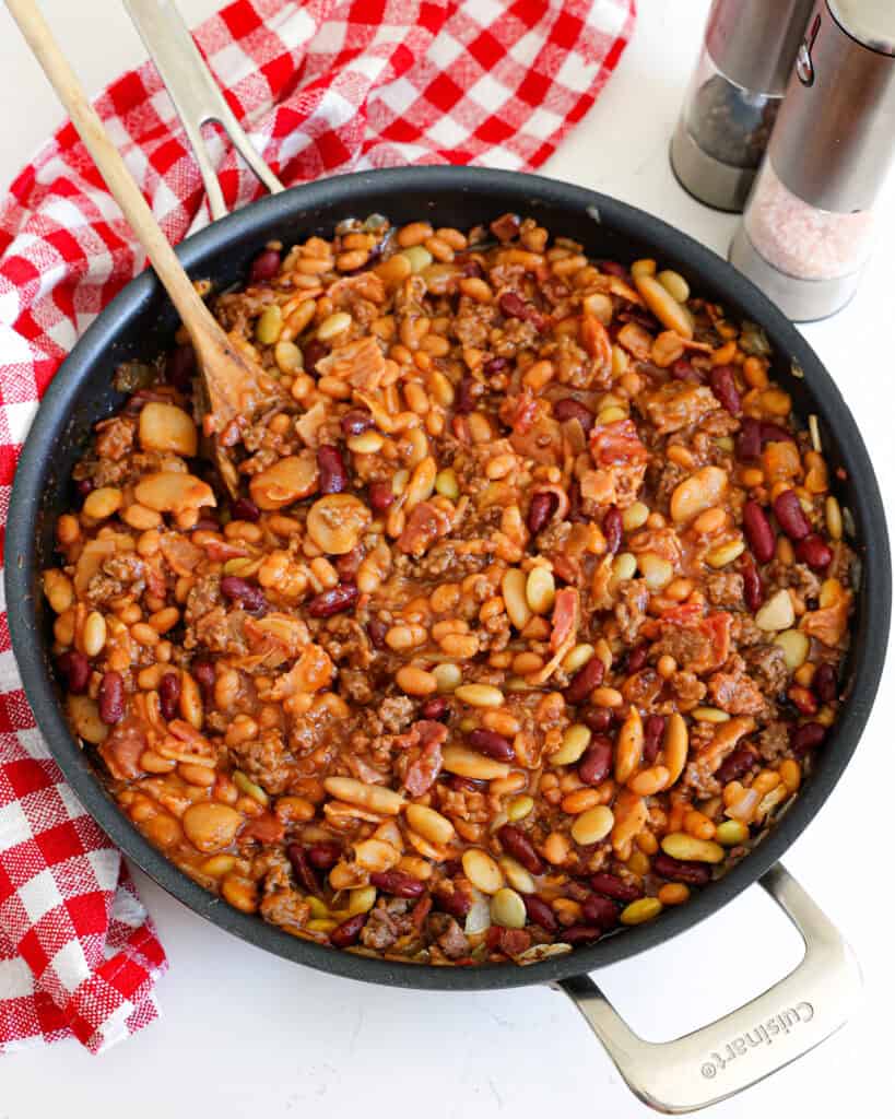 Overhead view of a large skillet full of calico beans with a salt and pepper shaker and a red and white checkered towel off to the side. 