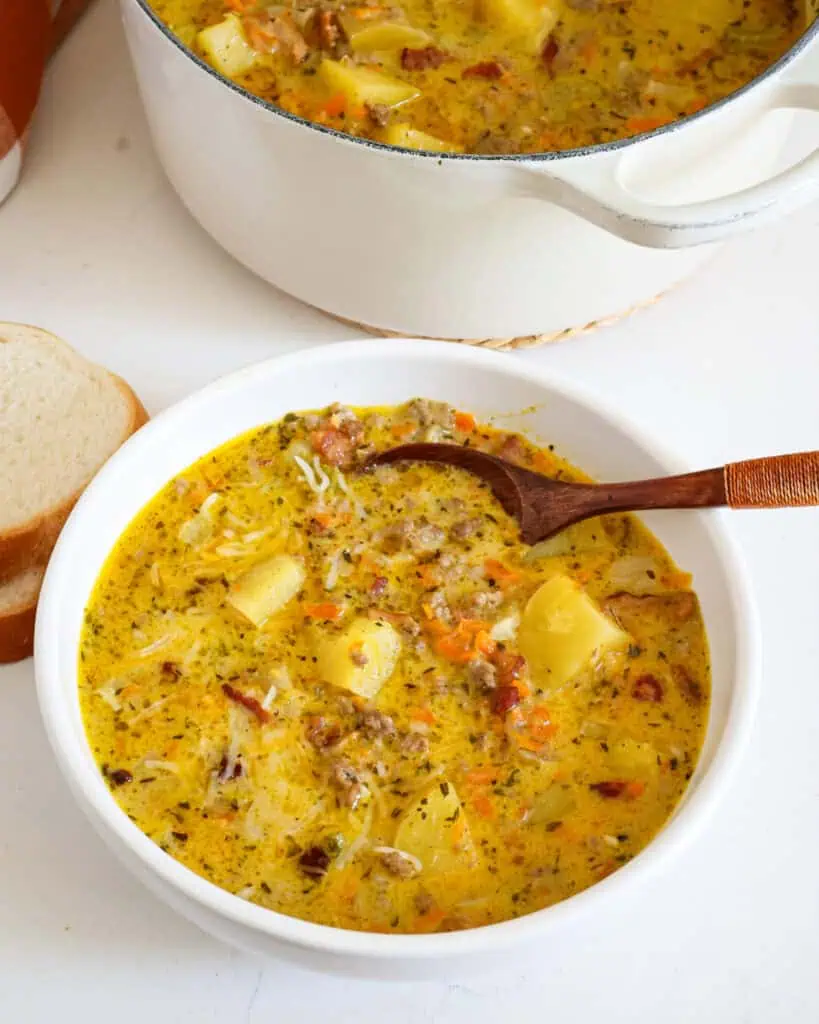 A bowl full of cheeseburger soup with a wooden spoon in it and twoe pieces of soft bread in the background. 