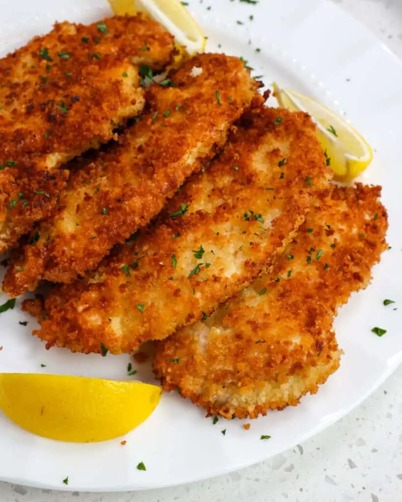 Multiple pieces of chicken schnitzel on a plate with lemon wedges. 