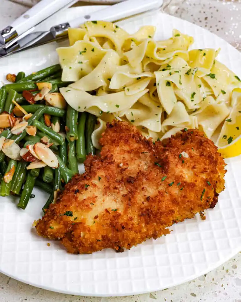 Chicken schnitzel on a plate with buttered noodles and green beans almondine. 
