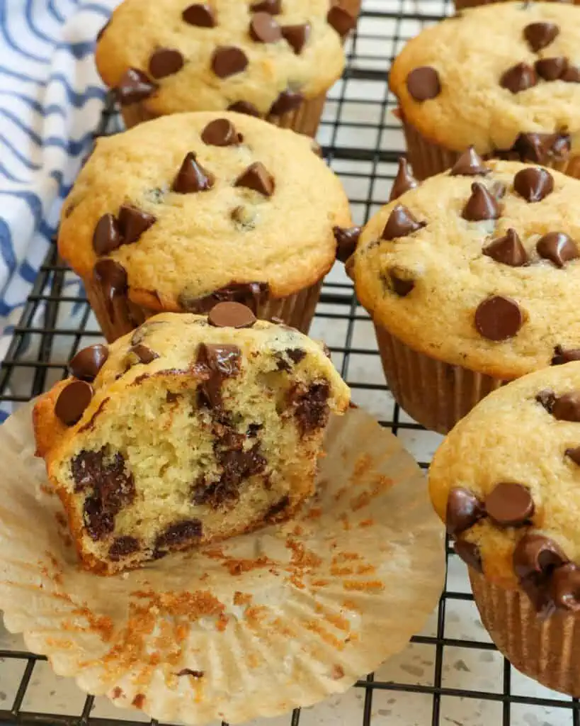 A chocolate chip muffin cut in half on a cooling rack with other whole chocolate chip muffins. 
