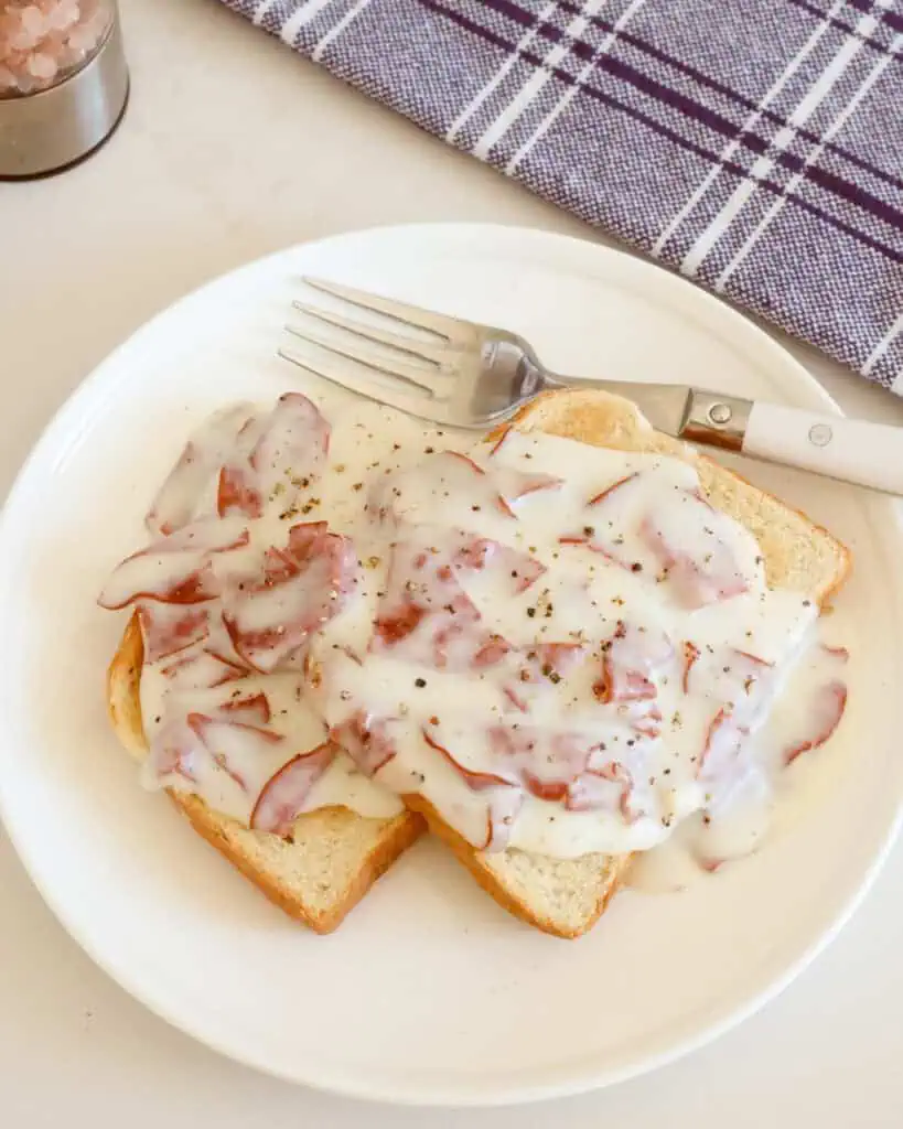 Two pieces of toast smothered in creamed chipped beef on a single serving plate with a fork. 