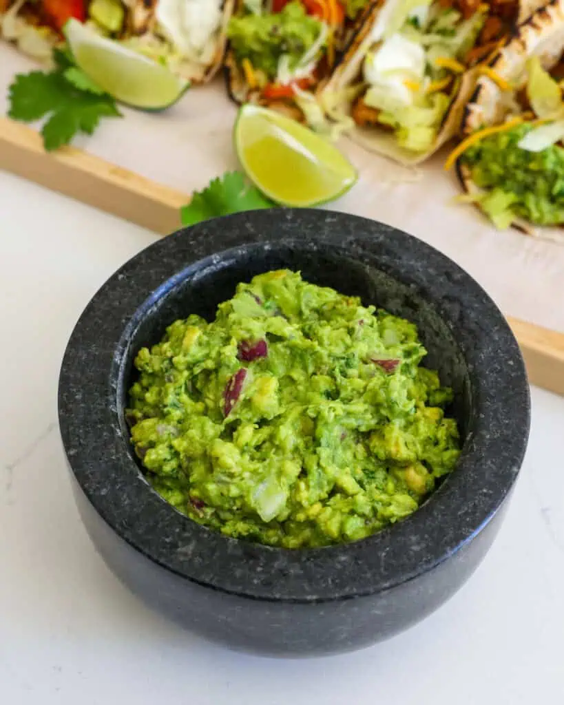 A mortar bowl full of guacamole in front of cutting board full of tacos. 