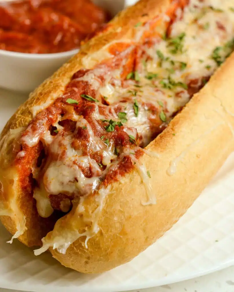 A single meatball sub on a plate with extra marinara on the side. 