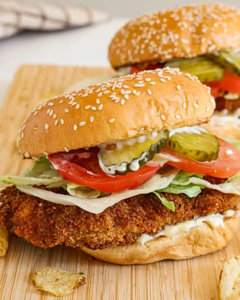 Two fried pork tenderloin sandwiches on a cutting board with potato chips. 