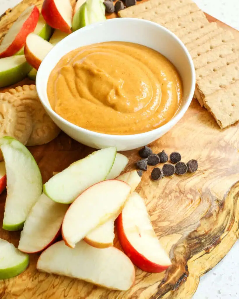 Pumpkin dip on a charcuterie board with red and green apple wedges, graham crakers, butter cookies, and chocolate chips. 