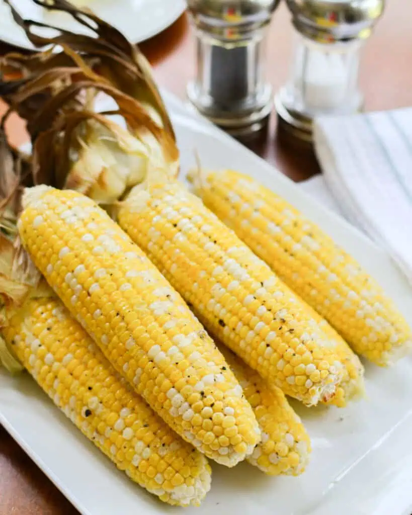 Six ears of buttered oven roasted corn on a plate with salt, black pepper, and butter in the background. 