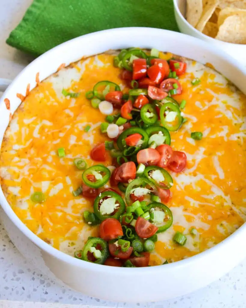 A large casserole dish filled with bean dip and topped with diced tomatoes, jalapeno rings, and sliced green onions. 