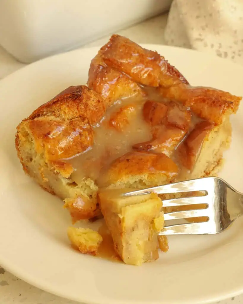 A piece of bread pudding on a plate with a fork with one bite on it. 