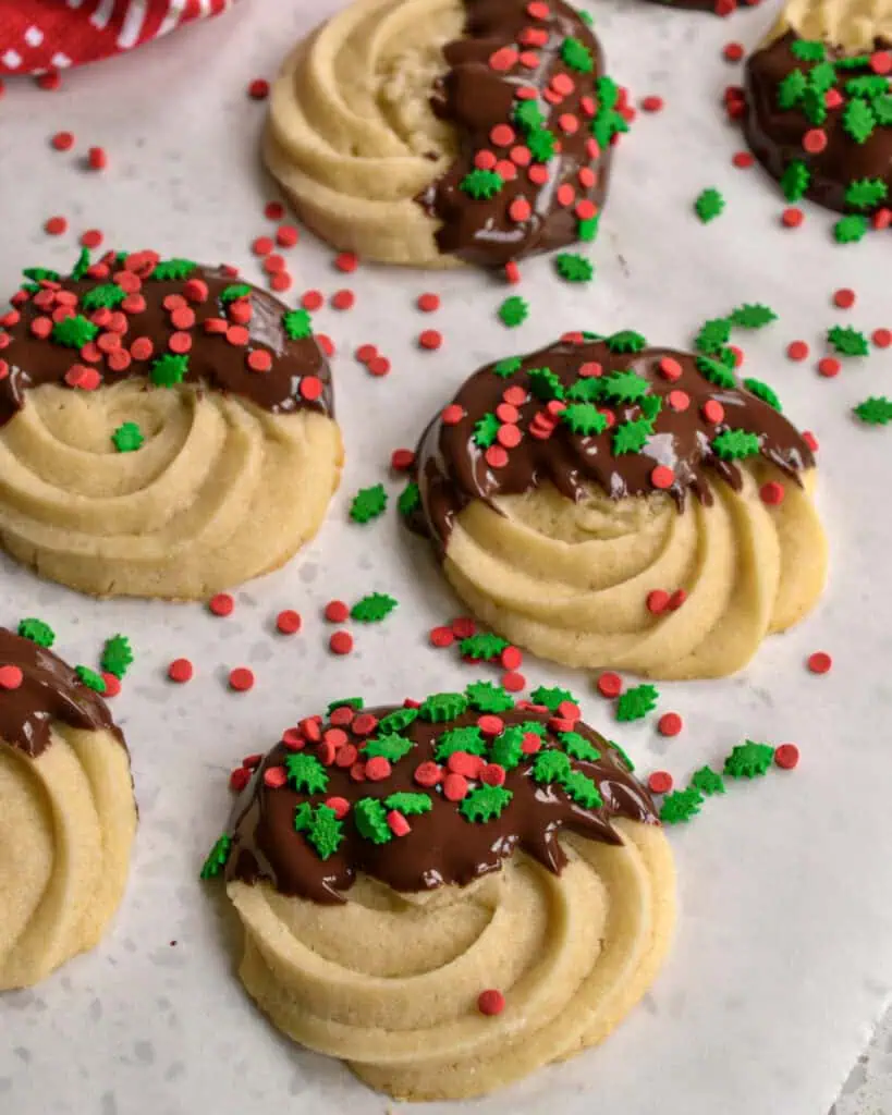 Butter cookies dipped in chocolate and sprinkled with Chrstmas sprinkles on a parchment covered baking sheet. 