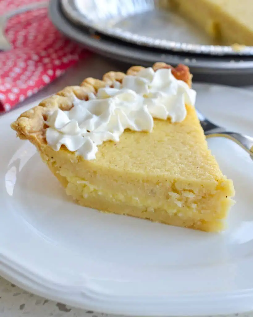 A slice of buttermilk pie topped with fresh whipped cream with more of the pie in the bacckground. 