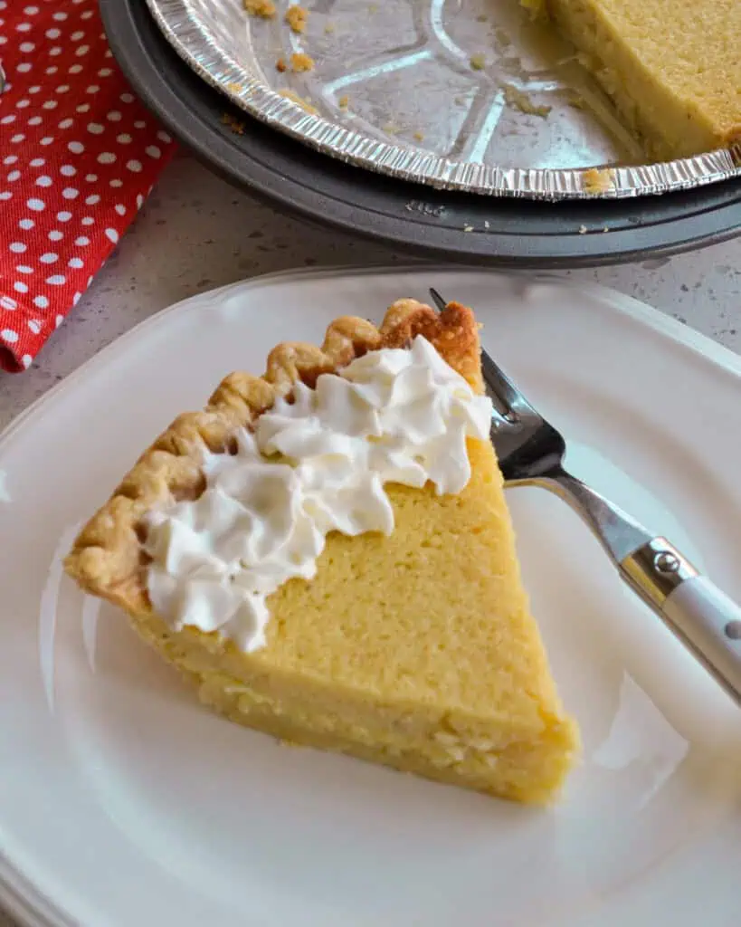 An overhead view of a sinlge slice of buttermilk pie with whipped cream on top on a single serving plate with a fork. 
