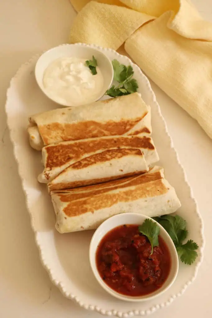 Three burritos on a platter with cilantro and a bowl of sour cream and a bowl of salsa. 