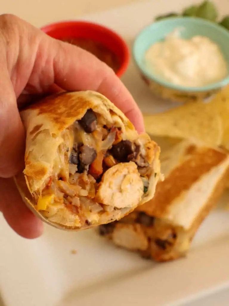 A hand holding a split chicken burrito with the other half of the burrito and chips in the background. 