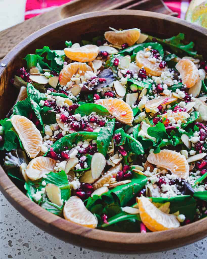 A large wooden bowl full of Christmas salad with almonds, mandarin oranges, feta cheese, and pomegranite seeds. 