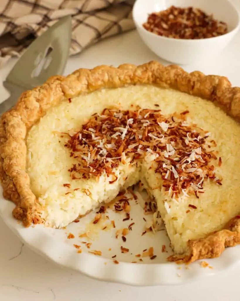 A whole coconut custard pie with one slice removed. With a pie server and a bowl of coconut in the background.