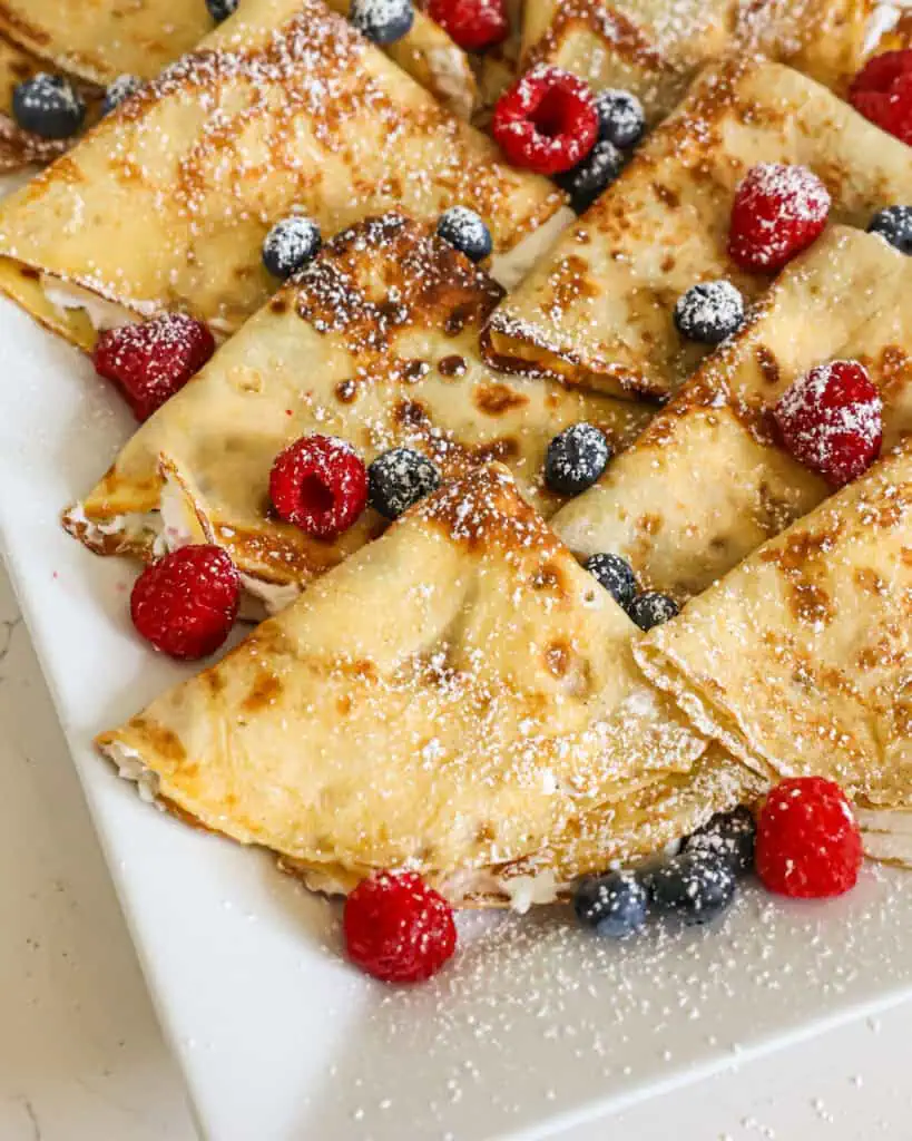 Whipped cream filled crepes on a platter with fresh raspberries and blueberries. 