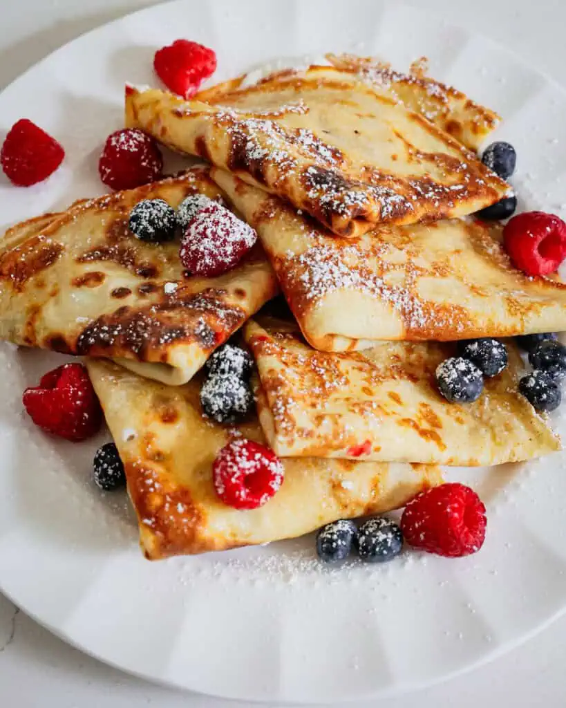 Whipped cream filled crepes sprinkled with powdered sugar on a plate with fresh blueberries and raspberres. 