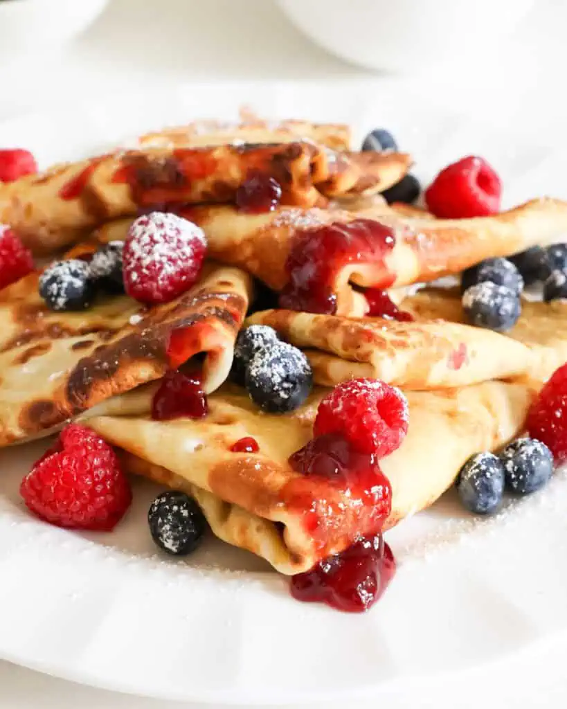 Folded stuffed crepes sprinkled with powdered sugar, raswpberry jam, an fresh fruit on a plate. 