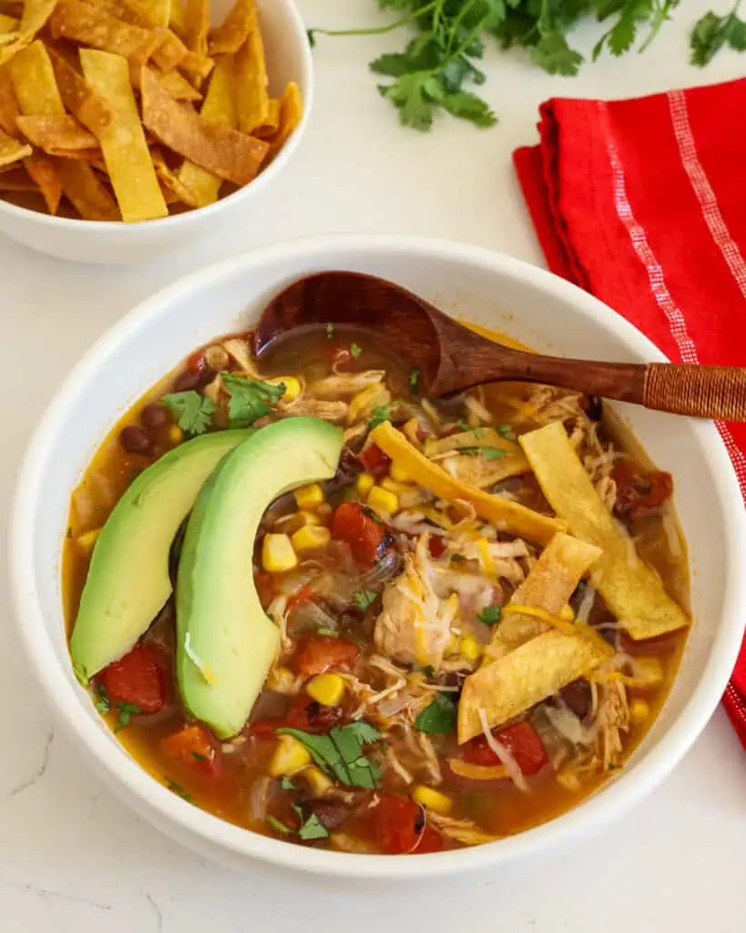 Crock pot chicken tortilla soup topped with sliced avocado, cheese, cilantro and fried tortilla strips in a bowl. 