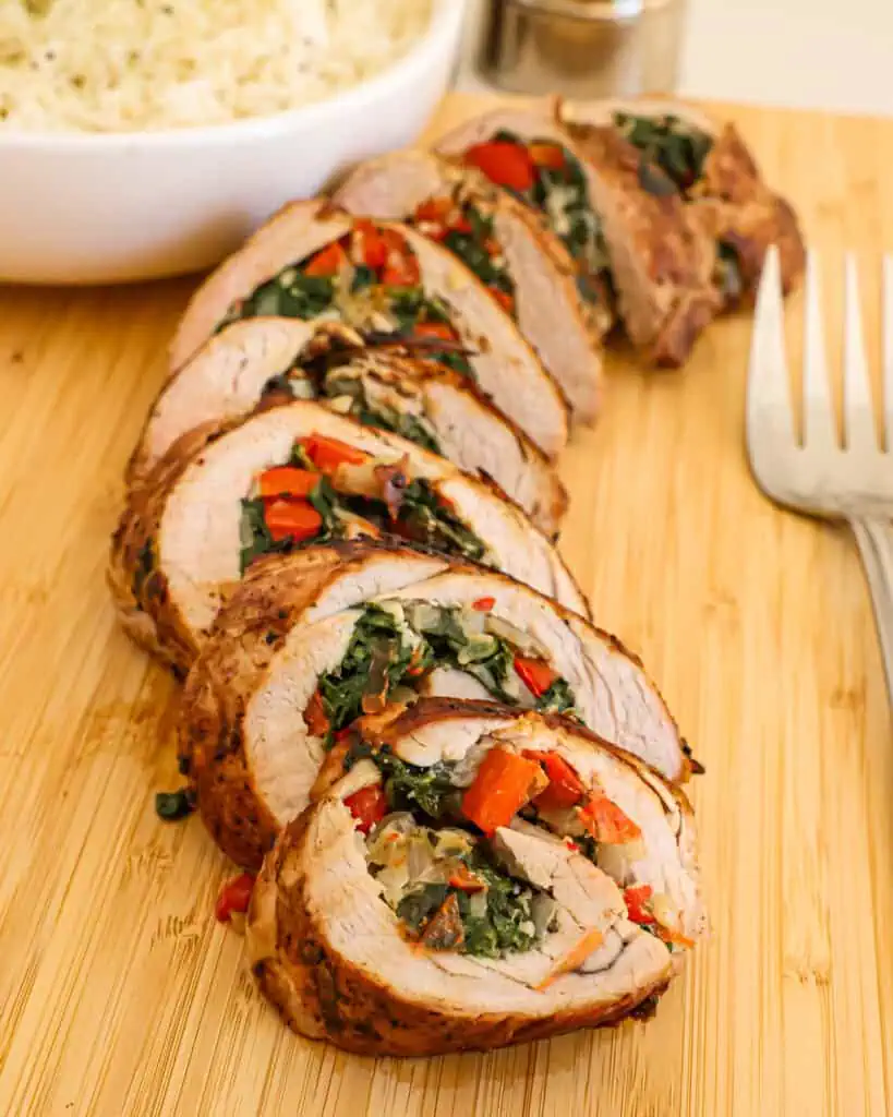 Sliced stuffed pork tenderloin on a wooden cutting board with bowl full of rice and a meat fork. 