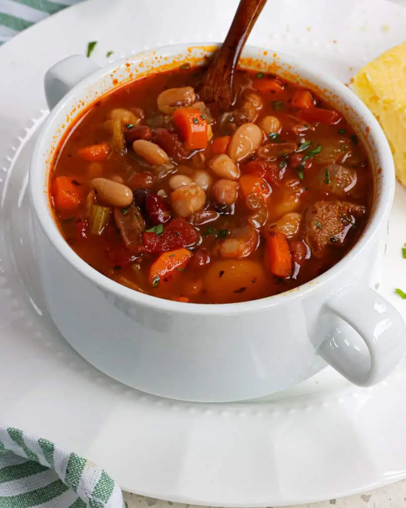 A soup crock full of 15 bean soup with  beans, lentils, onions, celery, carrots, and smoked sausage in a broth seasoned with smoked paprika, chili powder, and thyme.