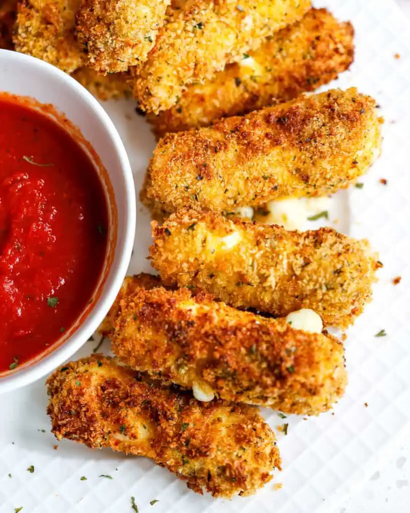 Air fried mozzarella sticks on a round plate with a bowl of marinara sauce. 