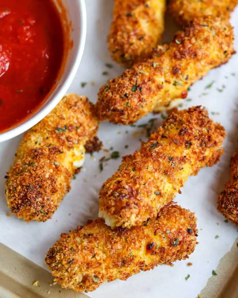 Air fryer mozzarrella sticks on a baking sheet with marinara sauce in a bowl. 