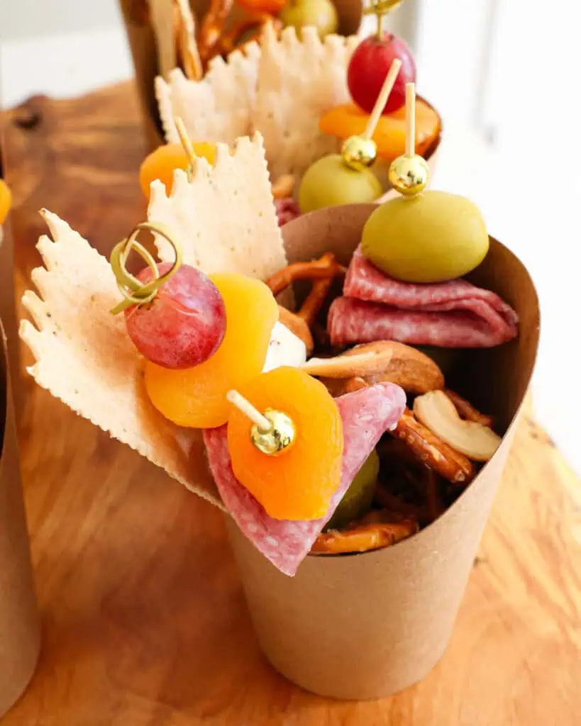 Charcuterie cup with pretzels, nuts, crackers, dried apricots, grapes, salami, and green olives.