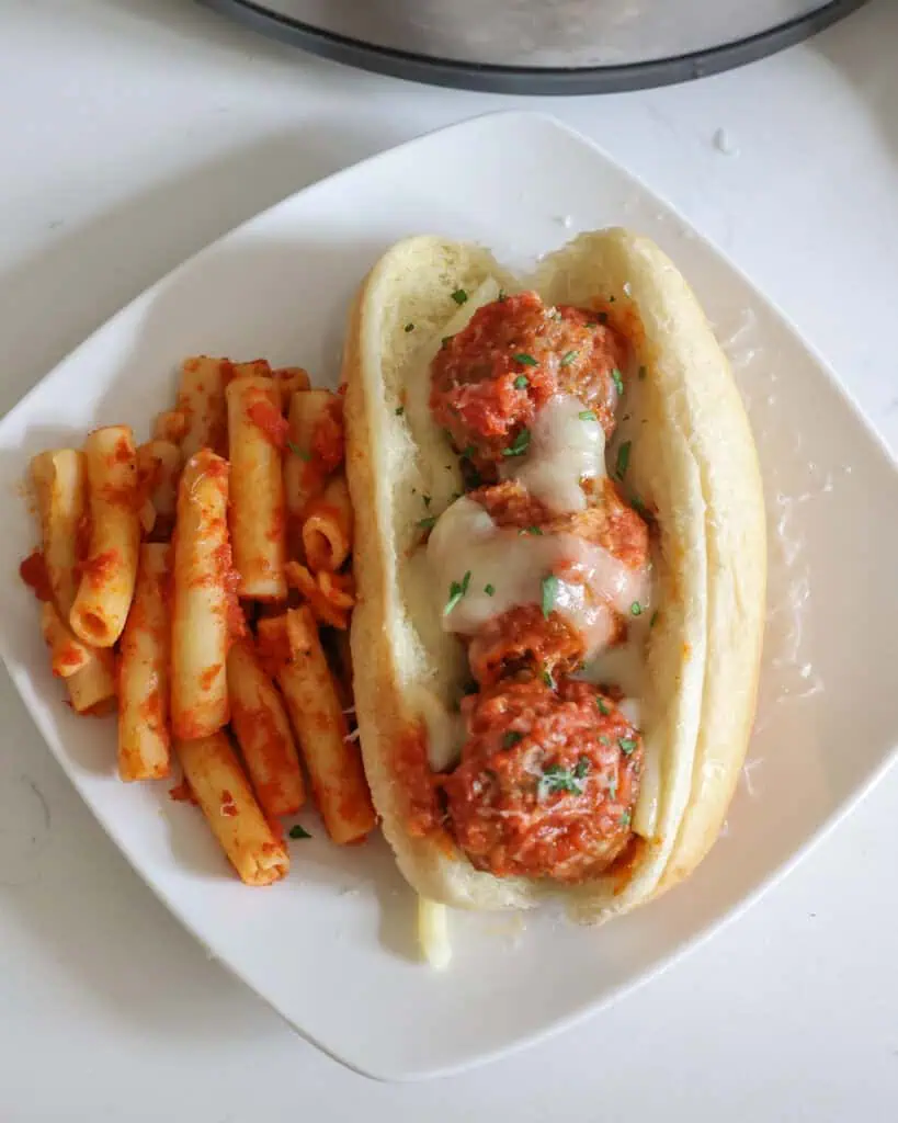 Crock pot meatballs with melted cheese over them in a toasted bun on a plate with a side of red pasta. 