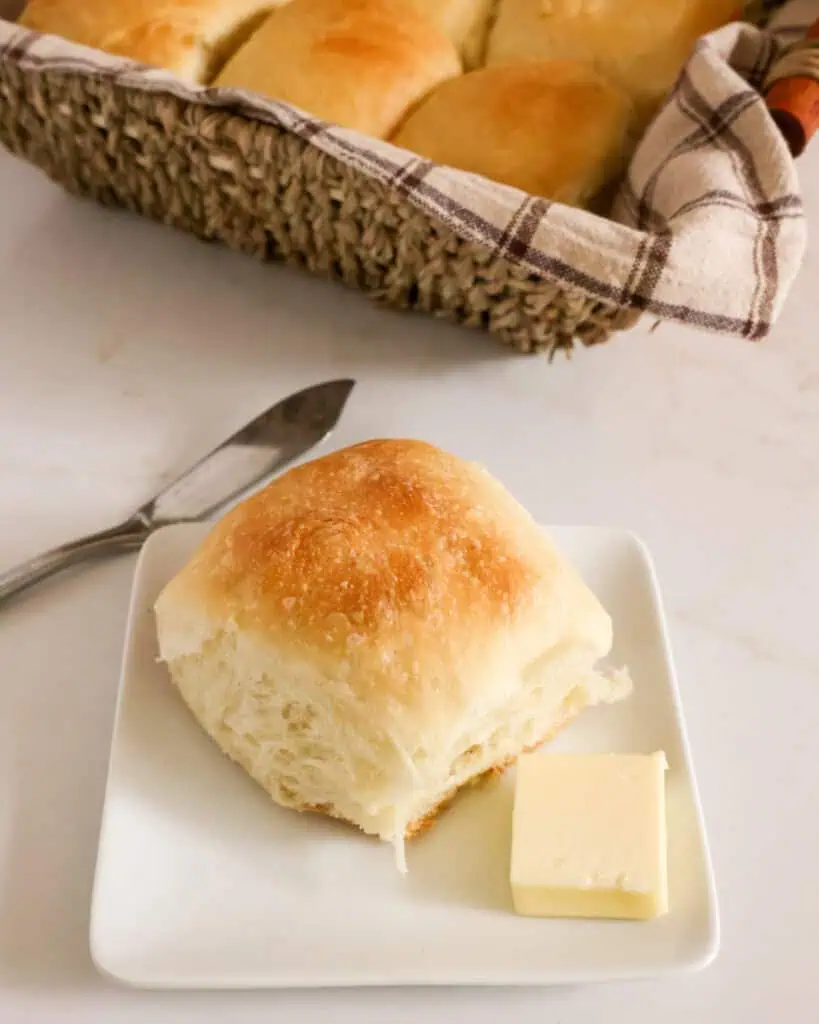 A homemade dinner roll on a plate with butter and a basket of dinner rolls in the background. 