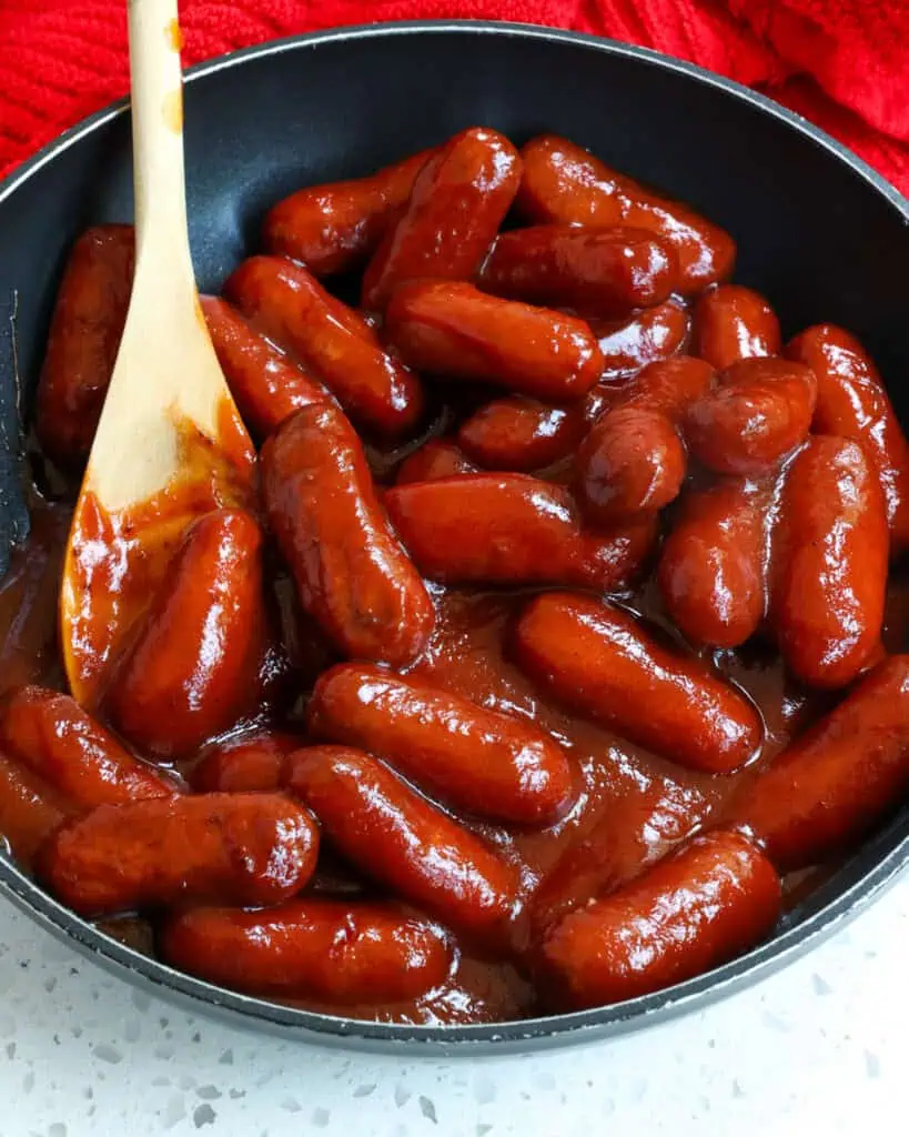 Little smokies in homemade sauce in a skillet with a wooden spoon. 