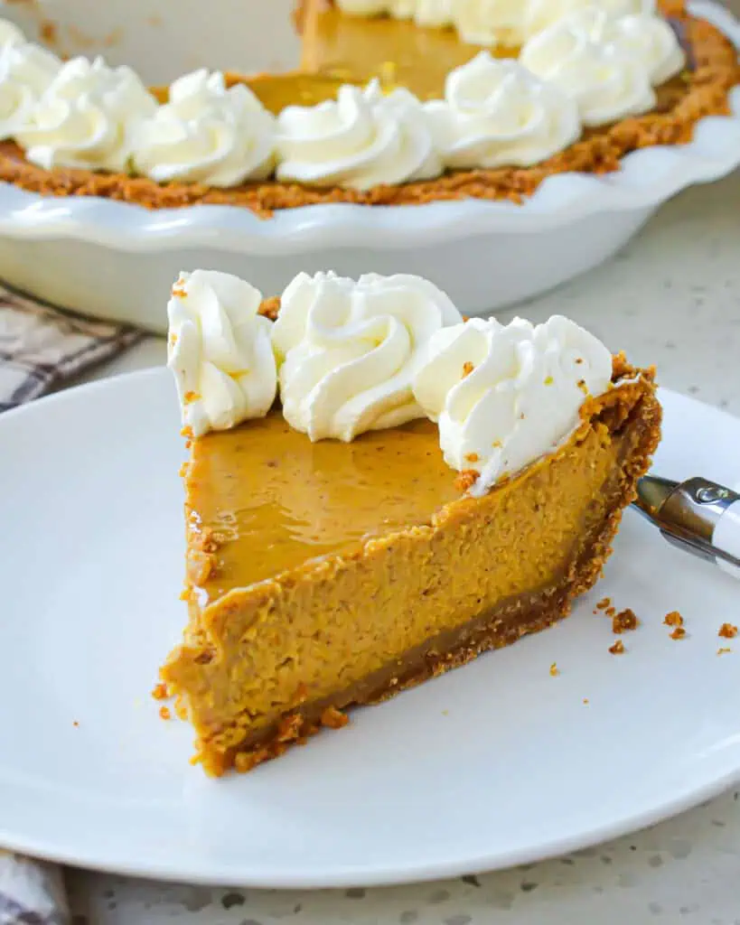 A slice of pumpkin pie with whipped cream on a plate with a fork and the rest of the pumpkin pie in the background. 