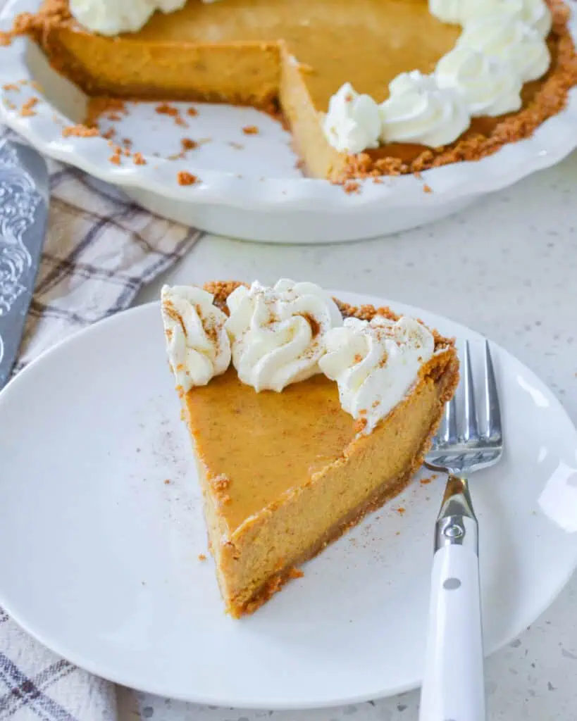 A slice of homemade pumpkin pie with whipped cream and the rest of the pumpkin pie in the bacckground. 