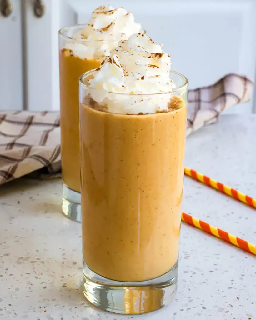 Two glasses full of pumpkin smoothie topped with whipped cream and cinnamon. 