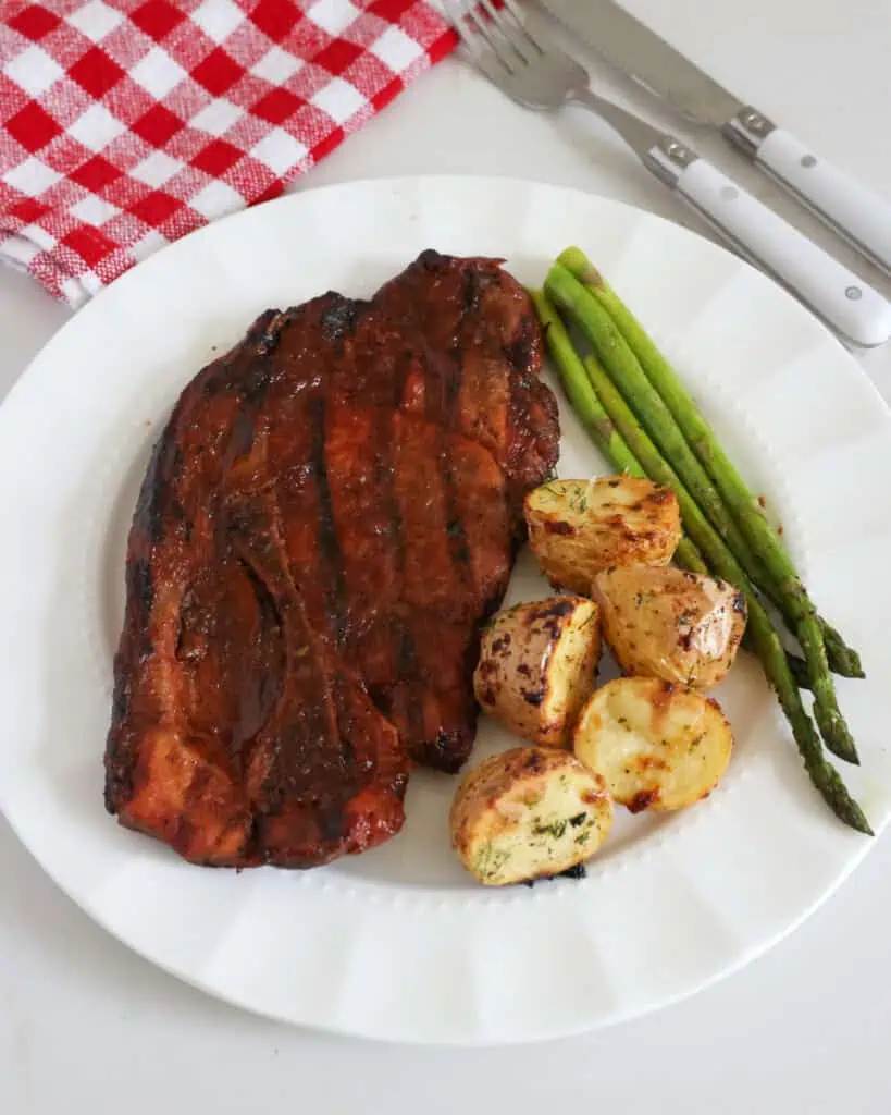 A pork steak with ranch potatoes and asparagus on a serving plate with a fork and knife in the background. 