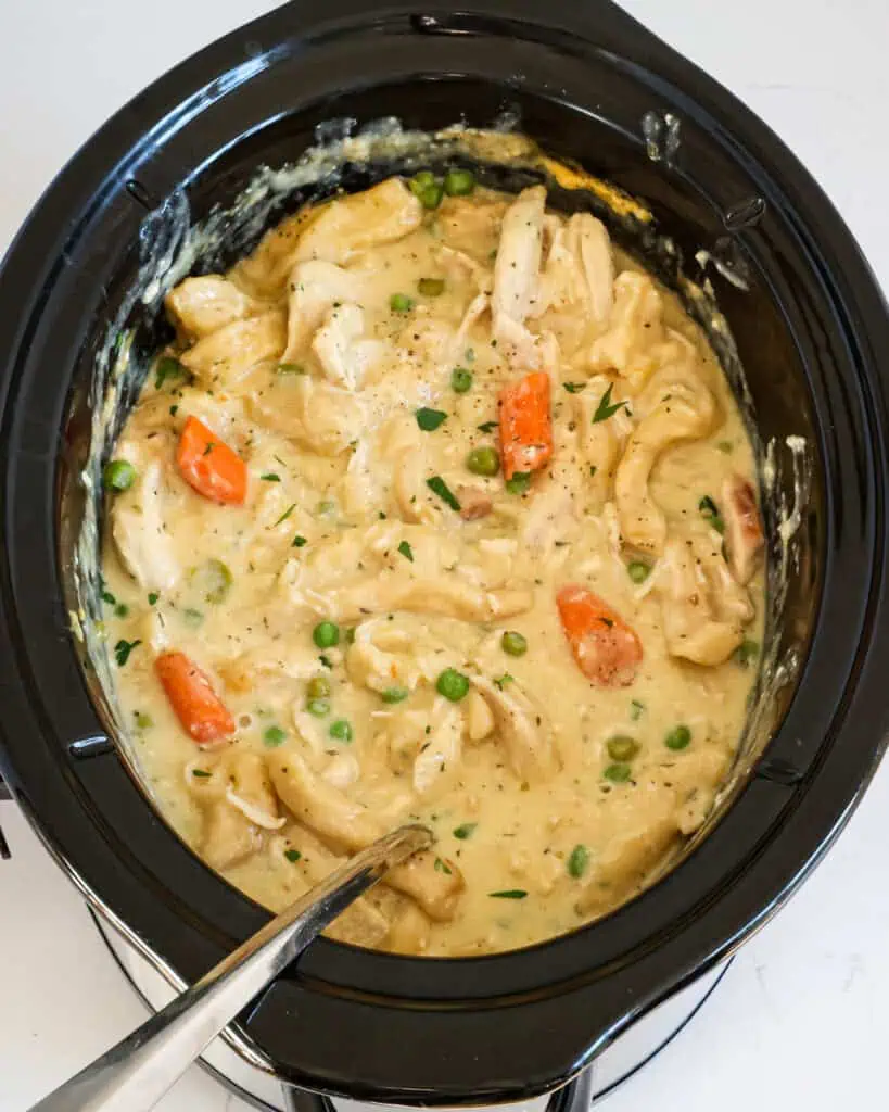 A crock pot full of chicken and dumplings with onions, peas, and carrots. 