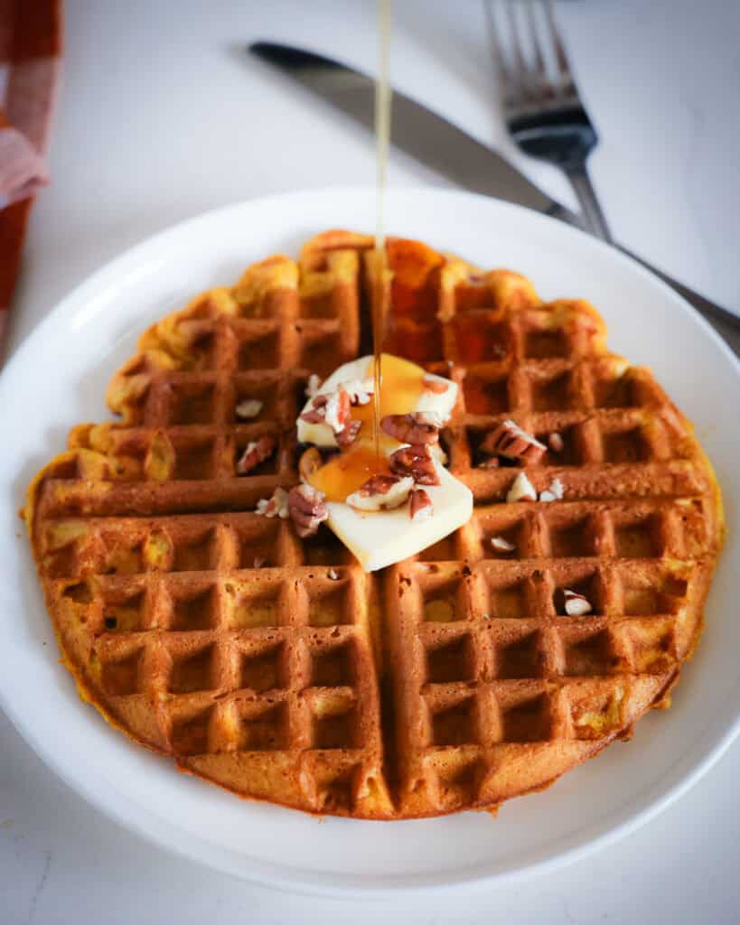 A pumpkin waffle topped with butter, pecans, and maple syrup on a single serving plate. 