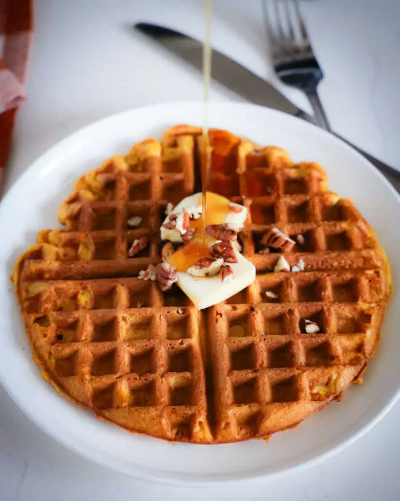 A pumpkin waffle topped with butter, pecans, and maple syrup on a single serving plate. 