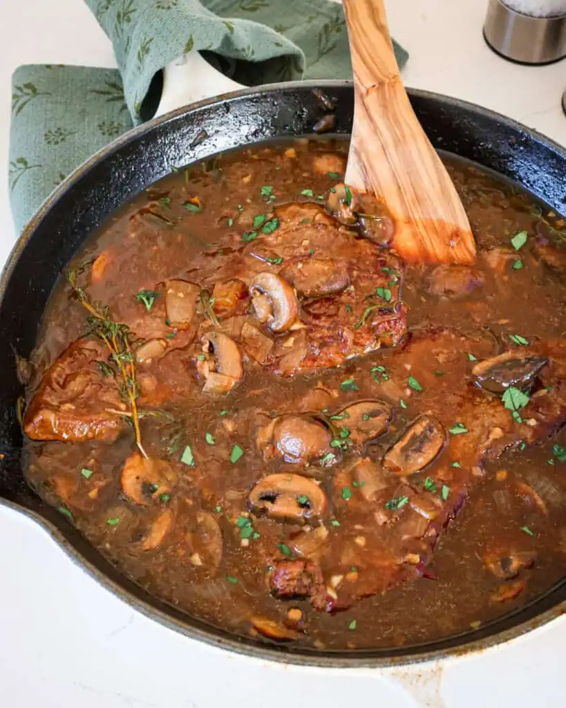 A cast iron skillet full of tender round steak smothred in gravy. There is a spatula in the pan and kitchen towel on the handle of the pan. 