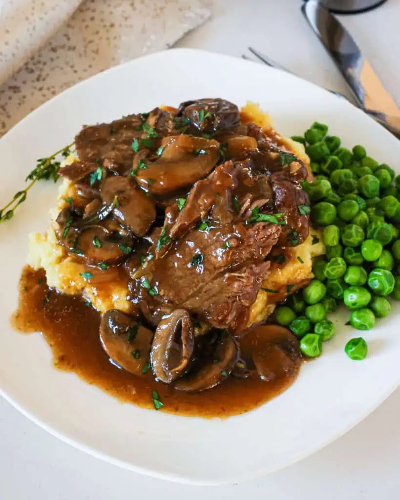 Round steak with gray and mushrooms over mashed potatoes with a side of baby peas on a single serving plate. 