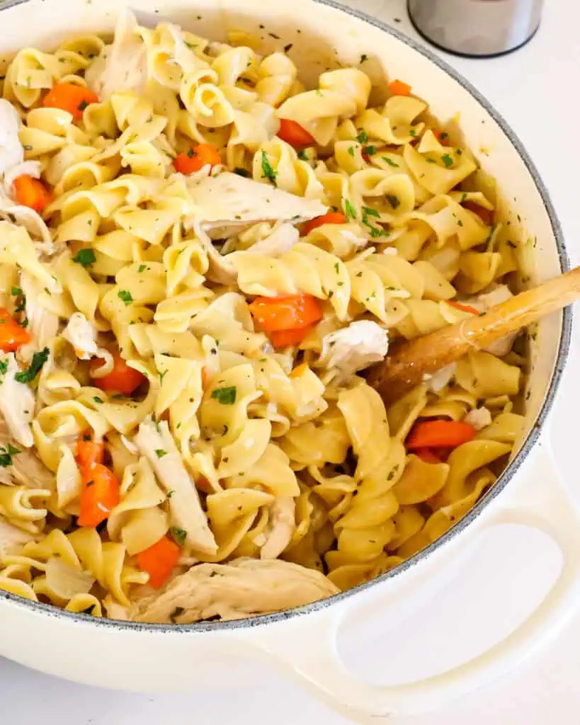 A close up view of chicken and noodles in a Dutch oven with a wooden spoon in it. 